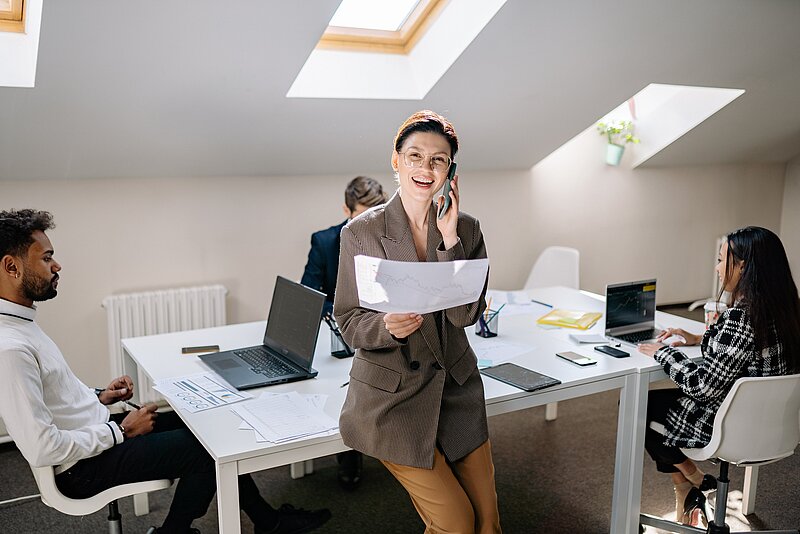 Eine Frau lehnt sich an einen Schreibtisch, hält ein Telefon an ihr Ohr und hält mit der anderen Hand eine ausgedruckte Grafik. An dem Tisch sitzen drei Personen und arbeiten an Laptops.