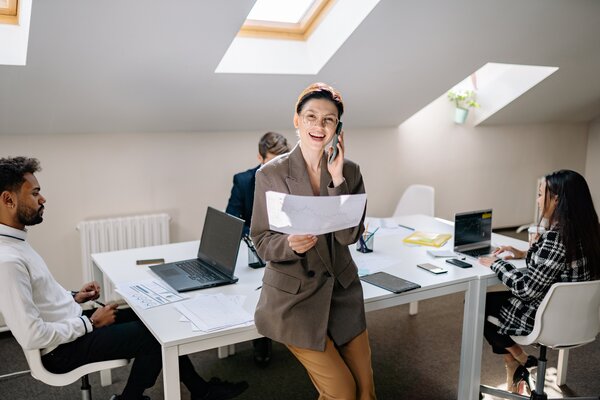 Eine Frau lehnt sich an einen Schreibtisch, hält ein Telefon an ihr Ohr und hält mit der anderen Hand eine ausgedruckte Grafik. An dem Tisch sitzen drei Personen und arbeiten an Laptops.